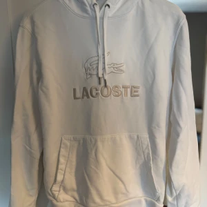 Vit hoodie från Lacoste med brodyr - Säljer en vit hoodie från Lacoste med broderad logga och krokodil på bröstet. Tröjan har huva med snörning och en stor magficka. Hoodien är i  storlek S för män. Använt ett par gånger. 