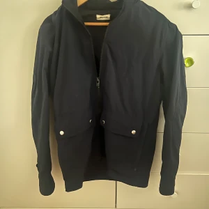 Svart vindjacka från Zadig Man - Svart vindjacka från Zadig Man med dragkedja framtill och två stora fickor med tryckknappar. Jackan har en enkel och stilren design, perfekt för blåsiga dagar. Långärmad och tillverkad i ett lätt material som känns smidigt att bära. Små fläckar som knappast syns. Frakt betalar köparen