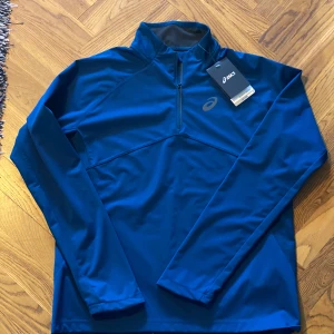 Blå träningshoodie från Asics M - Sportig blå hoodie från Asics med half zip och lång ärm. Perfekt för löpning eller träning, tillverkad i syntetmaterial som andas. Snyggt Asics-logga på bröstet och hög krage för extra komfort. Stilren och funktionell design.