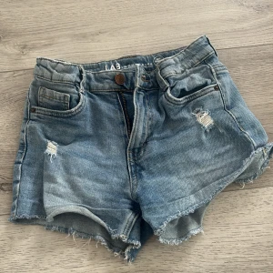 Blå jeansshorts från LAB Industries - Snygga blå jeansshorts från LAB Industries med fransiga kanter och slitna detaljer framtill. Klassisk femficksmodell med knapp och dragkedja. Perfekt för dig som gillar en chill och avslappnad stil på sommaren.
