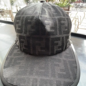 Svart keps från Fendi med logomönster - Säljer en svart keps från Fendi med det klassiska FF-logomönstret över hela kepsen. Kepsen har böjd skärm och justerbar rem baktill med Fendi Roma-broderi. Materialet är textil med en lyxig känsla och mönstret ger en riktigt exklusiv vibe.