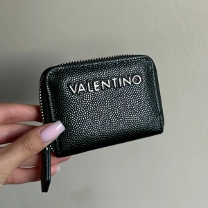 Valentino plånbok - Snygg svart plånbok från Valentino med silverfärgad logga framtill. Plånboken är i skinn med struktur och har dragkedja runtom. Perfekt storlek för kort och mynt, har ej haft så länge. köptes för ca 600kr säljes för 215kr 