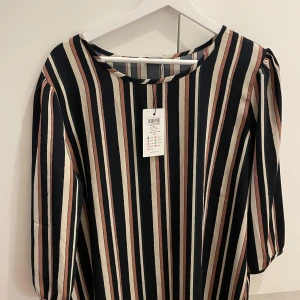 Randig klänning från Jacqueline de Yong - Snygg randig klänning från Jacqueline de Yong i storlek 42. Klänningen har långa ärmar och ett coolt mönster med vertikala ränder i marinblått, beige och rosa. Materialet är mjukt och lätt polyester, perfekt för dig som gillar stilrena och trendiga plagg.