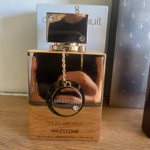 Club de Nuit Milestone parfym - Snygg och lyxig parfymflaska i guld från Armaf, Club de Nuit Milestone. Flaskan har en fyrkantig form med dekorativa stenar och en kedja med rund medaljong. Rymmer 105 ml Eau de Parfum. Perfekt för dig som gillar exklusiv design.