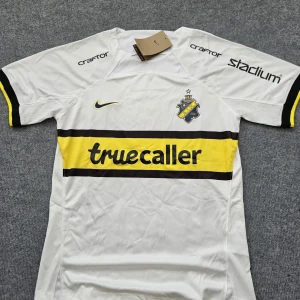 AIK vit bortatröja Nike S - AIK:s vita bortatröja från Nike i Dri-FIT-material. Tröjan har korta ärmar, svart och gul rand över bröstet med Truecaller-tryck, AIK-emblem och Nike-logga. Stadium, Craftor och SVEA-loggor på ärmar och rygg. Perfekt för dig som vill visa ditt AIK-stöd.