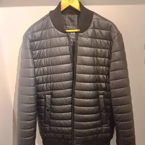 Svart quiltad bomberjacka DR MEN XL - Snygg svart quiltad bomberjacka från DR MEN i storlek XL. Jackan har horisontella sömmar, ribbade muddar och krage samt två fickor framtill. Materialet är syntet och ger en clean, modern look. Perfekt för dig som vill ha en stilren jacka med edge.