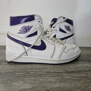 Nike Air Jordan 1 High vit/lila - Säljer ett par Nike Air Jordan 1 High i vitt och lila skinn. Klassisk high-top siluett med lila swoosh, detaljer och Air Jordan-logga på sidan. Skorna har snörning och perforerad tå för extra ventilation. Perfekta för dig som vill sticka ut med en ikonisk sneaker.