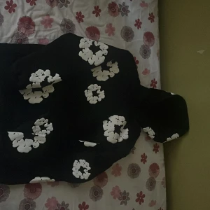 Svart hoodie med vita blommor Denim Tears - Svart hoodie från Denim Tears med stora vita blommor tryckta över hela tröjan. Klassisk huva och lång ärm, perfekt för dig som gillar streetwear och vill sticka ut. Mjuk bomullskänsla och relaxed fit. Snygg detalj med blommönster som ger en unik vibe.