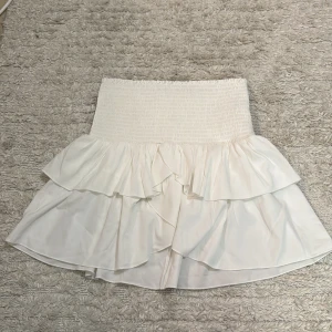 Vit volangkjol från Neo Noir - Supersöt vit kortkjol från Neo Noir med bred smock i midjan och dubbla volanger. Kjolen har en flowy och luftig känsla, perfekt för att skapa en fräsch och trendig look. Materialet är mjukt och lätt, vilket gör den skön att bära under varma dagar.