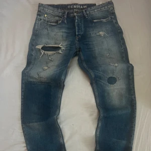 Denham jeans - Säljer ett par blå jeans från Denham med coola slitningar, patchar och hål för en riktigt edgy look. Jeansen har klassisk femficksdesign, tydliga slitdetaljer och broderad sax på bakfickan. Perfekta för dig som gillar streetstyle och vill sticka ut. Jeansen är helt oanvända.