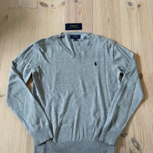 Grå v-ringad tröja från Polo Ralph Lauren - Tvär skön grå v-ringad tröja från Polo Ralph Lauren med klassisk broderad logga på bröstet. Tröjan har långa ärmar och ribbade muddar vid ärmslut och nederkant. Perfekt för lager-på-lager och stilrena outfits. PRIS KAN DISKUTERAS!! Den passar ungefär runt 158! Använd endast 2 gånger!