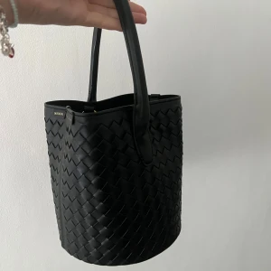 Svart bucket bag från Don Donna - Snygg svart bucket bag från Don Donna med flätad design och rund form. Väskan har både handtag och axelrem i matchande svart, samt detaljer i guld. Tillverkad i skinnimitation och rymmer allt du behöver till vardagen. Skriv för egna bilder. Kan gå ner i snabb affär. Andvänd 1 gång 