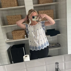 Vit blus med blå broderi och volang - Supersöt vit blus med blått broderat mönster och volangkant. Blusen har korta ärmar och rund halsringning, samt broderade detaljer både på ärmar och längs nederkanten. Perfekt för en fräsch och somrig look.