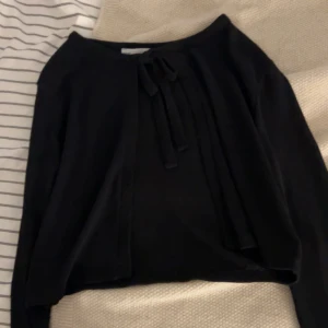 Svart långärmad tröja med knyt - Svart långärmad tröja med snygg knytning framtill vid halsen. Enkel och stilren design med rak passform, perfekt att matcha med jeans eller kjol. Mjuk och skön känsla, passar dig som gillar minimalistisk stil.