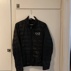 Svart pufferjacka från EA7 Armani - Snygg svart pufferjacka från EA7 Emporio Armani med diskret glansig finish och logga på bröstet. Jackan har dragkedja framtill, ståkrage och quiltad design. Perfekt för dig som vill ha en stilren och sportig look under kyliga dagar. Ett litet hål under märket finns men inget man tänker på jätte mycket.