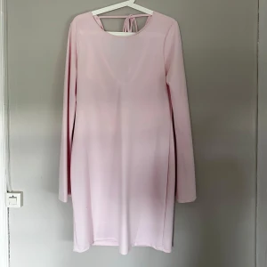 Ljusrosa långärmad klänning med draperad rygg - Säljer en ljusrosa klänning med lång ärm och snygg draperad rygg. Klänningen har en kort längd och knytning i nacken som ger en extra detalj. Materialet känns mjukt och har ett fint fall. Perfekt för dig som gillar stilrena och feminina plagg.