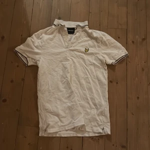 Vit pikétröja från Lyle & Scott - Klassisk vit pikétröja från Lyle & Scott med korta ärmar och gul logga på bröstet. Tröjan har krage med knappar och svarta detaljer på krage och ärmslut. Tillverkad i bomull, perfekt för en clean och sportig stil.