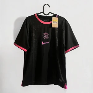 Svart PSG x Nike fotbollströja med rosa detaljer - Snygg svart PSG fotbollströja från Nike med rosa och lila detaljer på krage och ärmslut. Tröjan har klubbmärke och Nike-logga framtill, samt diskret mönster i tyget. Tillverkad i lätt och ventilerande material, perfekt för match eller träning.