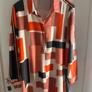 Färgstark mönstrad skjorta med knappar - Snygg skjorta med grafiskt mönster i rött, svart, beige och vitt. Skjortan har lång ärm med knappdetaljer och klassisk krage. Materialet känns lätt och luftigt, perfekt för dig som gillar att sticka ut med färg och form.