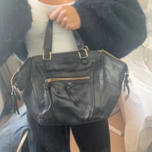 Svart handväska i skinn med dragkedjor - Snygg svart handväska i skinn med guldiga metalldetaljer och flera dragkedjor. Väskan har två rejäla handtag och en praktisk ytterficka framtill. Perfekt storlek för att få plats med allt du behöver och passar till många olika outfits.