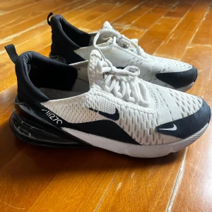 Nike Air Max 270 vita och svarta sneakers - Nike Air Max 270 sneakers i vitt och svart med stor synlig Air-enhet i hälen. Ovandelen är i ventilerande mesh med svarta detaljer och tydliga Nike-loggor. Skorna har snörning och en sportig, modern look som passar perfekt till streetwear.