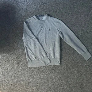 Grå sweatshirt från Polo Ralph Lauren - Klassisk grå sweatshirt från Polo Ralph Lauren med broderad logga på bröstet. Tröjan har rund halsringning, ribbade muddar och är tillverkad i mjuk bomull. Perfekt för en chill och stilren look.