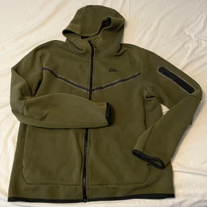 Nike Tech fleece olivgrön  - Nike hoodie i olivgrönt med hel dragkedja och svart logga på bröstet. Tröjan har huva, lång ärm och en snygg detalj med svart rand över bröstet samt ficka med dragkedja på ena ärmen. Perfekt för en chill och avslappnad stil.(Pris kan diskuteras) Använd fåtals gånger.