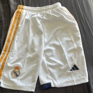 Real Madrid vita fotbollsshorts Adidas - Snygga vita Real Madrid shorts med gula ränder längs sidorna och klubbens emblem broderat på ena benet. De har elastisk midja och mörkblå detaljer vid bensluten. Perfekta för fotboll eller träning, tillverkade i lätt polyester.