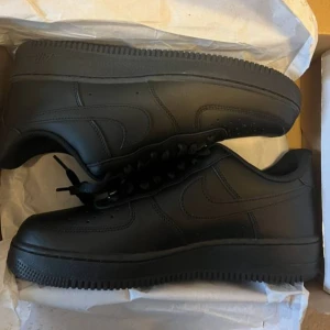 Svarta Nike Air Force 1 sneakers - Säljer ett par helsvarta Nike Air Force 1 sneakers i klassisk low-modell. Skorna är gjorda i skinn med matchande svarta snören och sula. Perfekta för dig som gillar en clean och stilren look. Ikonisk Nike-logga på sidan.