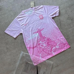 Rosa Brasil fotbollströja Linited edition - Riktigt stilren och unikt designad Brasilien-tröja i rosa! Kombinationen av färgen, loggan och den ikoniska Jesusstatyn gör den här tröjan till ett måste i garderoben.  Perfekt som present till dig själv, din partner eller varför inte köra en matchande outfit? Passa på att köpa din innan den blir viral, för det här är en tröja som garanterat kommer att väcka uppmärksamhet!