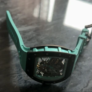 Klocka - Säljer en riktigt snygg klocka med turkost silikonarmband och svart boett. Klockan har ett skeletturverk där du ser kugghjulen och mekaniken genom glaset både fram och bak. Rektangulär form och coola detaljer i silver och turkos.