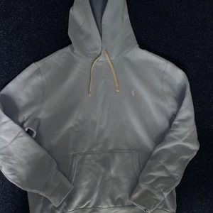 Ljusblå hoodie från Polo Ralph Lauren - Snygg ljusblå hoodie från Polo Ralph Lauren med klassisk huva och beige snörning. Tröjan har en stor magficka och det ikoniska lilla Polo-logot på bröstet. Perfekt för chill dagar och enkel att matcha med jeans eller joggers.