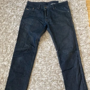 Diesel Darron mörkblå jeans W33 L32 - Snygga mörkblå jeans från Diesel, modell Darron. Regular slim-tapered passform med klassiska fem fickor och diskreta sömmar. Jeansen har orangea detaljer vid bälteshällorna och är tillverkade i slitstarkt denim. Perfekt för dig som gillar stilrena och moderna jeans.