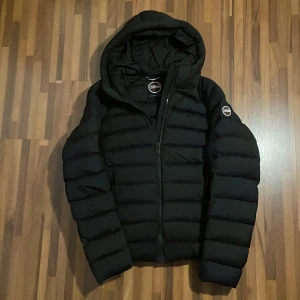 Svart Colmar pufferjacka med huva - Svart pufferjacka från Colmar med huva och två dragkedjeförsedda fickor. Jackan har en quiltad design och tydlig logga på ärmen. Perfekt nu till vintern.