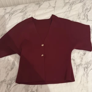 Vinröd blus med guldfärgade knappar - Vinröd blus med v-ringning och två snygga guldfärgade knappar framtill. Modellen är croppad med korta ärmar och har en stilren, loose passform. Perfekt för dig som gillar minimalistisk design och vill ha en blus som sticker ut med sin djupa färg och coola detaljer. Min mammas gamla ❤️