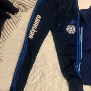 Adidas Djurgården träningsbyxor blå - Mörkblå träningsbyxor från Adidas med Djurgården-tryck och Volkswagen-logga på ena benet. Byxorna har blå ränder längs sidorna och snörning i midjan. Perfekta för fotbollsträning eller chill.