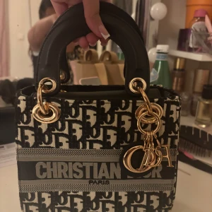 monogram handväska - Säljer en ikonisk handväska  med svart och beige monogrammönster, guldfärgade metalldetaljer och rejäla svarta handtag. Väskan har tydlig logga framtill och klassiska berlocker. Perfekt storlek för det viktigaste och riktigt snygg statement piece.