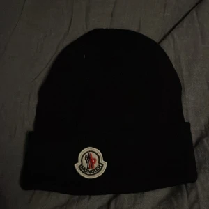 Svart Moncler mössa i ull - Svart mössa från Moncler med klassisk uppvikt kant och broderad logotyp framtill. Tillverkad i mjuk ull som håller dig varm under kyliga dagar. Perfekt accessoar för att lyfta din vinterstil.