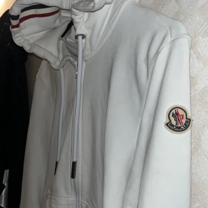 Vit hoodie från Moncler med huva - Säljer en stilren vit hoodie från Moncler med dragkedja framtill och klassisk huva med röd och marinblå rand. Tröjan har långa ärmar, fickor och Moncler-logga på ärmen. Perfekt för dig som gillar exklusiva streetwear-plagg.