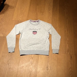 Grå sweatshirt från GANT med logga - Snygg grå sweatshirt från GANT med broderad logga och sköld på bröstet. Tröjan har rund hals. Strorleken är 15 years/170cm.