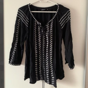 Svart och vit blus med snörning KappAhl - Svart blus från KappAhl i storlek XS med vita broderade mönster längs ärmar och framtill. Blusen har snörning vid halsen och trekvartsärmar. Lätt och luftig känsla, perfekt för en chill och snygg look.