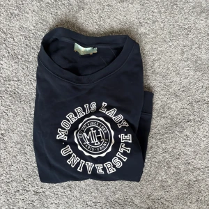 Mörkblå sweatshirt från Morris Lady - Snygg mörkblå sweatshirt från Morris Lady med vit universitetsinspirerad print framtill. Tröjan har rund halsringning och långa ärmar, perfekt för en chill och stilren look. Tillverkad i mjuk bomull som känns skön mot huden.