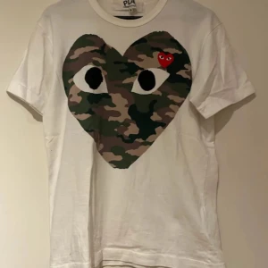 Vit Comme des Garçons Play t-shirt - Vit t-shirt från Comme des Garçons Play med stort hjärta i camouflage och ögon på bröstet, samt ett litet rött hjärta. Klassisk passform och rund hals. Perfekt statement-plagg för dig som gillar streetstyle.