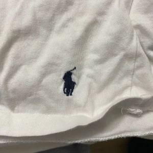 Vit t-shirt från Polo Ralph Lauren - Vit t-shirt från Polo Ralph Lauren i mjuk bomull. Klassisk design med korta ärmar och rund hals. Diskret mörkblå broderad logga med polospelare på nedre delen. Perfekt för en clean och chill stil.