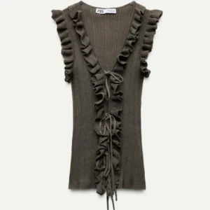 Volang topp zara  - Supersnygg brun ribbad topp från Zara med volanger längs v-ringningen och axlarna. Toppen är ärmlös och har knyt framtill som ger en extra cool detalj. Perfekt för dig som gillar unika plagg med lite edge och romantisk vibe.
