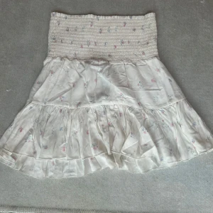 Vit blommig volangkjol från 157 - Supersöt kort kjol från 157 i vitt med små blommor i rosa och blått. Kjolen har smock i midjan och volang nertill som ger en flowy känsla. Perfekt för varma dagar och enkel att matcha med en crop top eller t-shirt.