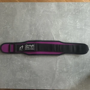 Träningsbälte Gorilla Wear svart/lila - Säljer ett brett träningsbälte från Gorilla Wear i svart och lila med justerbart kardborrespänne. Bältet har en tydlig logga med gorilla och texten 'I DARE YOU' på insidan. Perfekt för gymmet när du vill ha extra stöd vid tunga lyft.