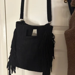 Svart axelväska med fransar i mocka - Cool maje liknande axelväska i mocka med långa fransar på sidorna och fyrkantigt handtag. Väskan har en bred axelrem och ett rymligt innerfack med dragkedja för småsaker. Perfekt för dig som behöver en fin väska till skolan eller annat💕