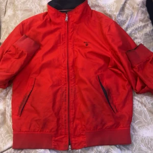 Röd vindjacka från Gant - Riktigt snygg röd vindjacka från Gant med svart foder och diskret logga på bröstet. Jackan har dragkedja framtill, ståkrage och två fickor med svarta detaljer. Perfekt för dig som vill ha en sportig och stilren look.