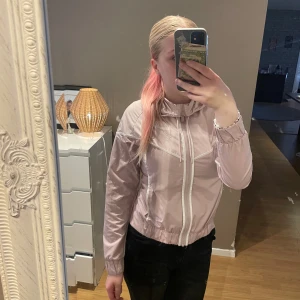 Ljusrosa vindjacka med dragkedja - Säljer en snygg ljusrosa vindjacka med hel dragkedja framtill och elastisk midja. Jackan har huva med snörning och vita detaljer längs dragkedjan och fickorna. Perfekt för dig som gillar en sportig och clean stil.